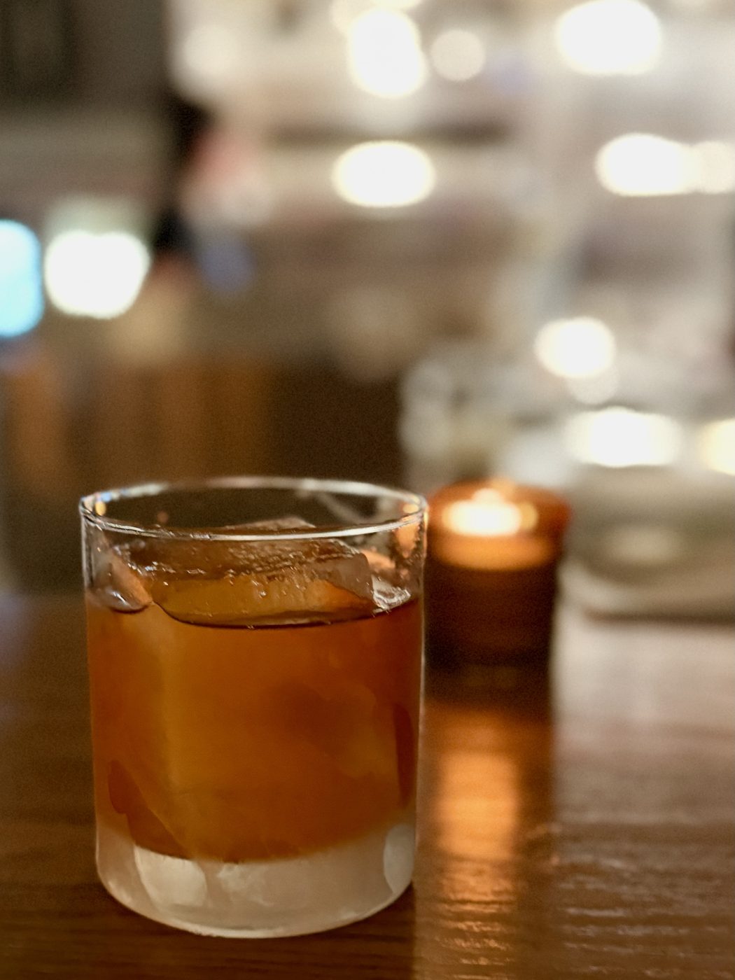 An amber cocktail over a single large ice cube in a heavy tumbler, a lit candle and the blurred lights of the dining room behind it