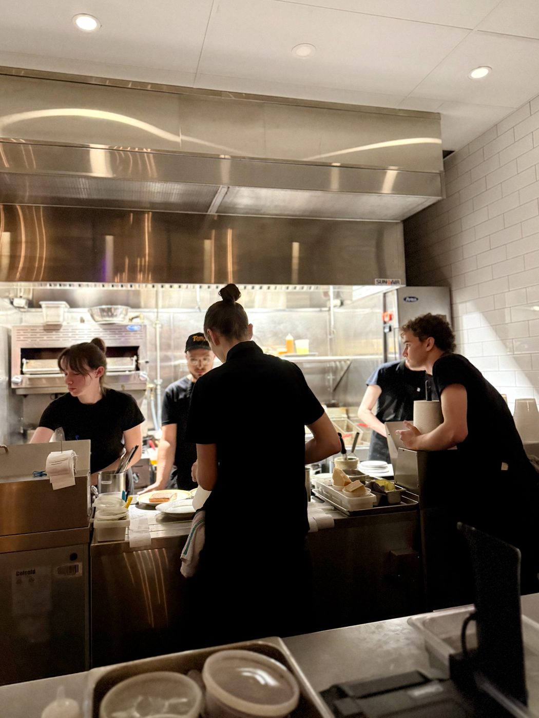 An open kitchen line mid-service under a stainless steel extraction hood, chefs in black working between the stove and the counter, plates waiting to go out