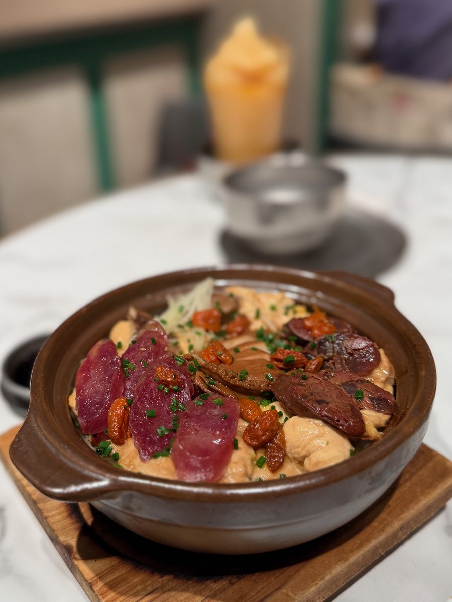 An open clay pot of rice topped with abalone, translucent slices of cured pork, sausage, goji berries and chives