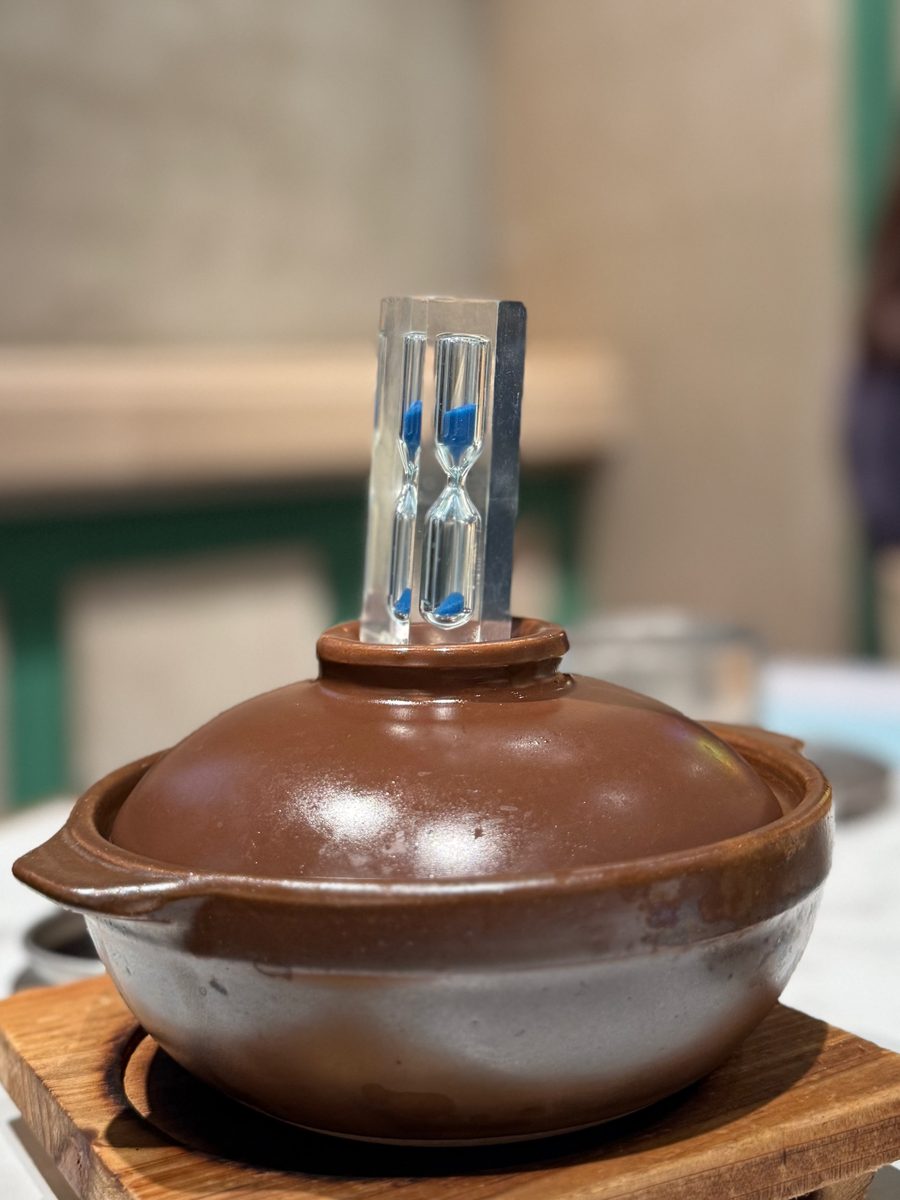 A lidded brown clay pot on a wooden board with a small two-tube sand timer set into the hole in the lid