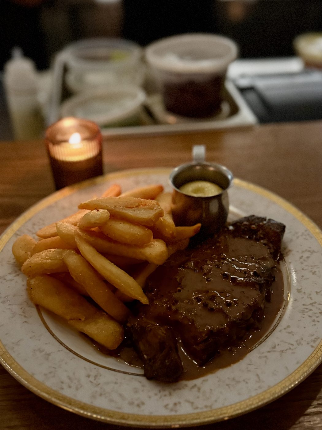 A seared steak under a glossy peppercorn glaze beside a pile of thick-cut fries and a small silver pot of pale aioli, on a gold-rimmed plate lit by a candle