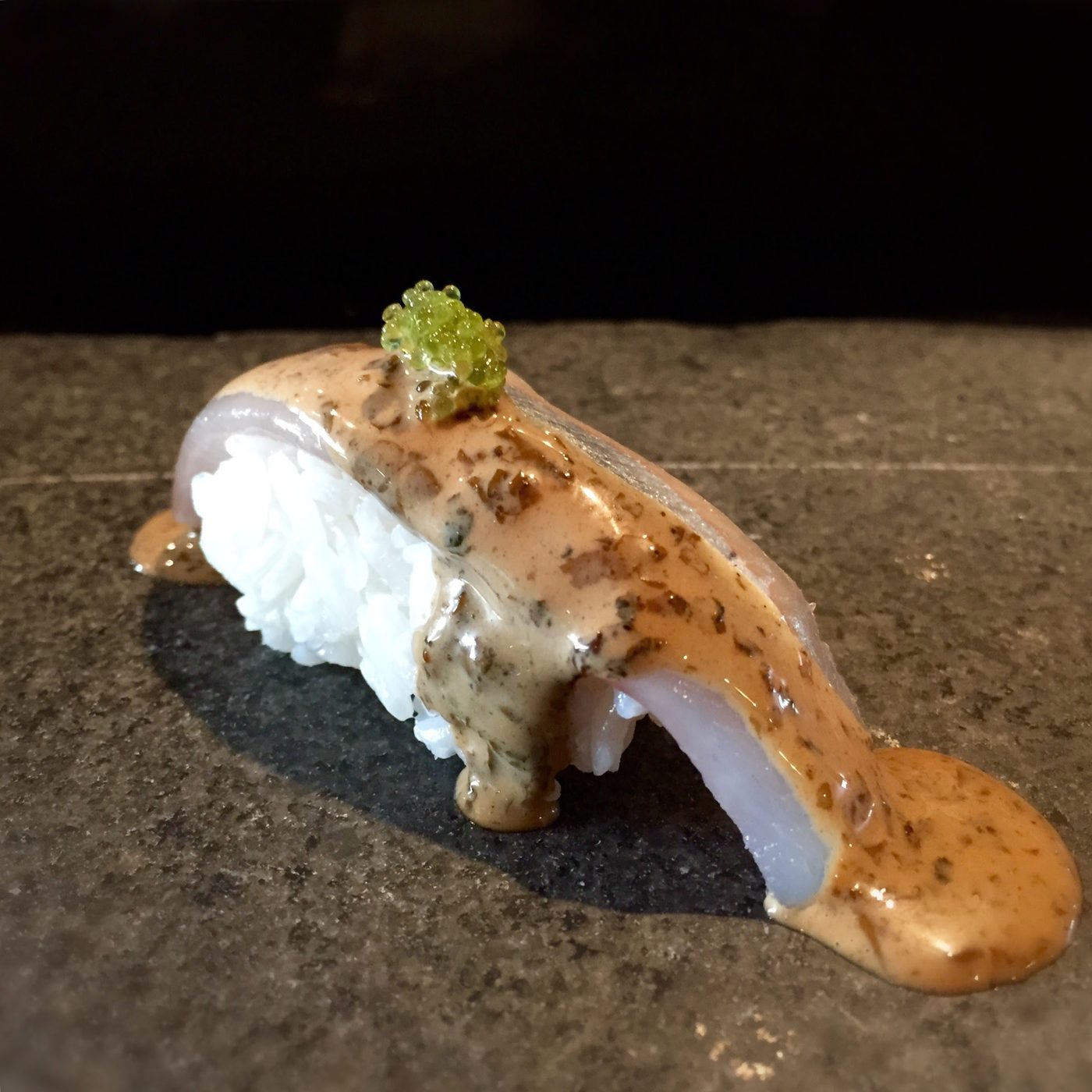 White fish nigiri under a heavy flecked nori emulsion with bright green wasabi tobiko, the sauce running onto a dark stone plate.