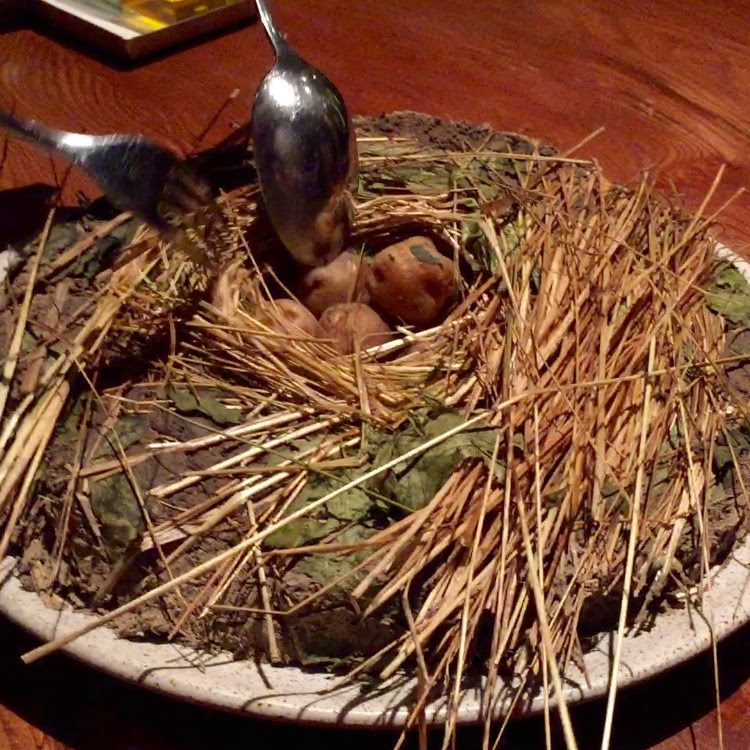 An Andean huatia opened at the table: straw pulled back from a bed of earth and scorched leaves, potatoes exposed in the ash, two spoons alongside.