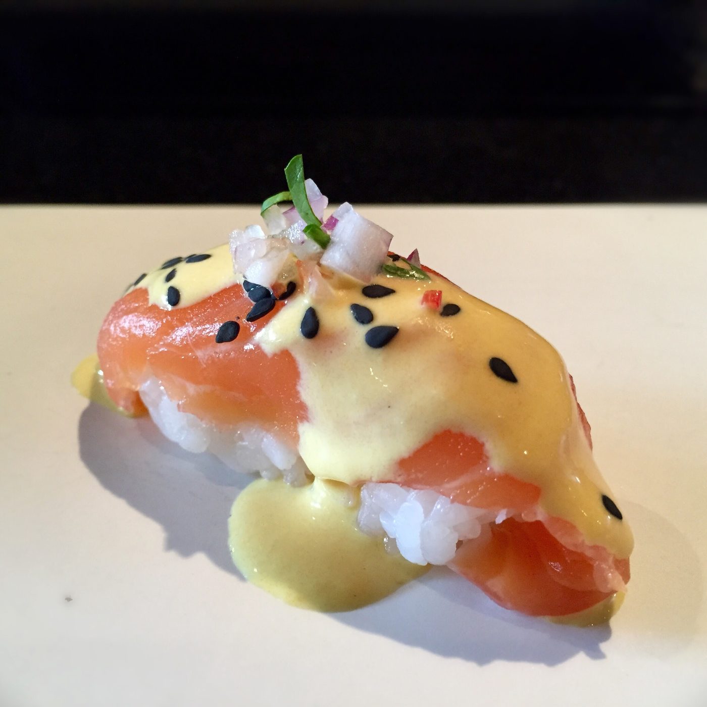 A salmon nigiri under yellow aji amarillo sauce, scattered with black sesame and diced red onion.