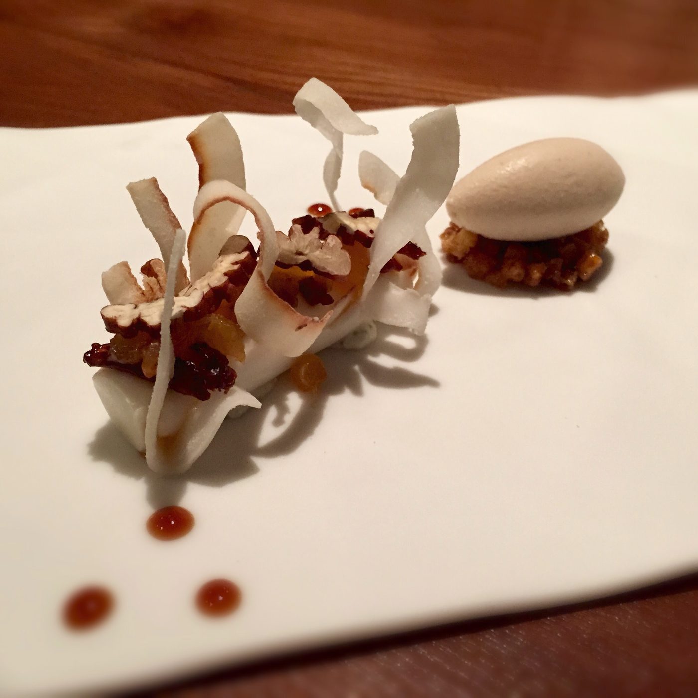 Wide coconut shavings standing off a white plate with pecans and raisins, a quenelle of ice cream on a nut crumble and three caramel dots below.