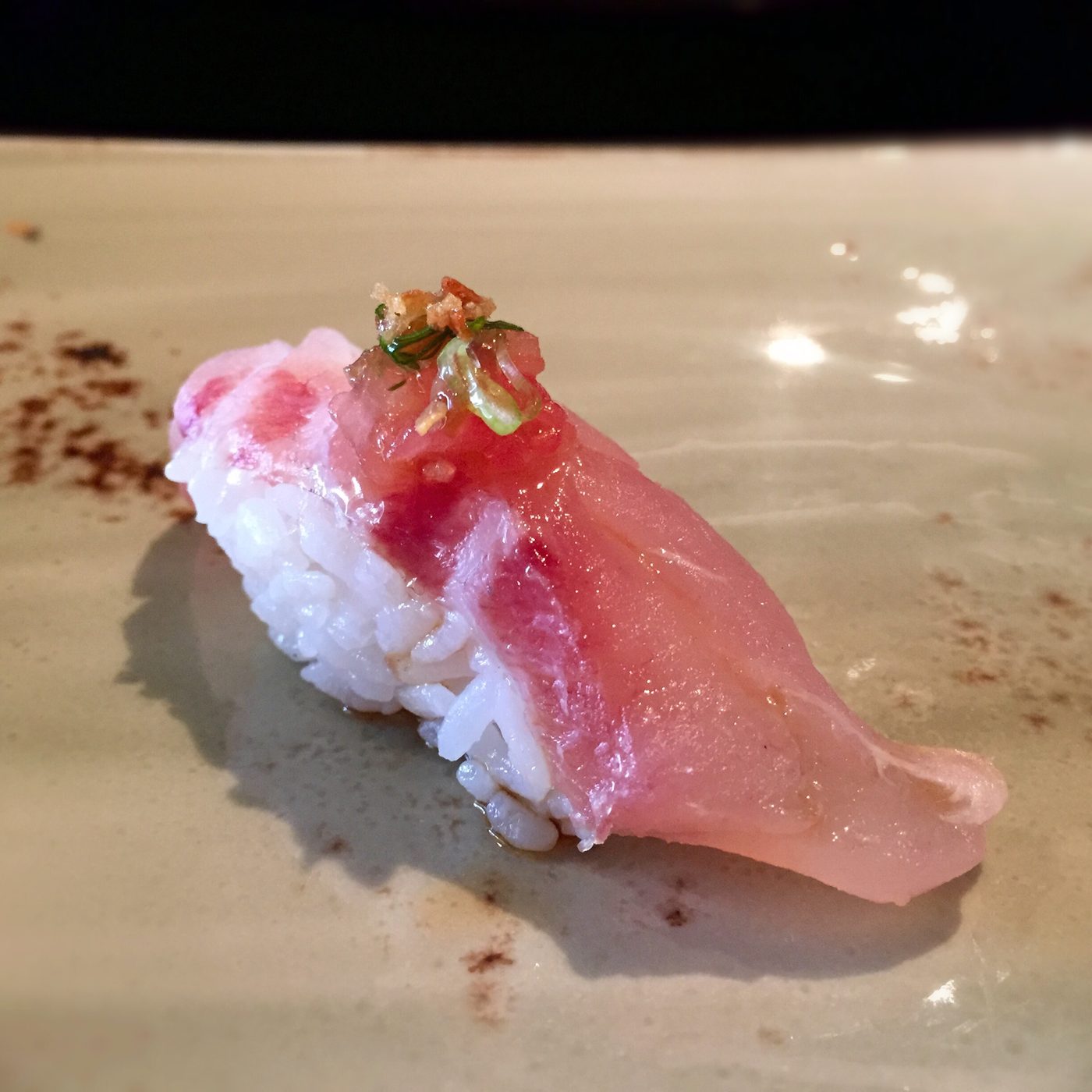 A nigiri of pale pink fish with a seam of dark red through it, finished with green scallion threads and crisp fried shreds.