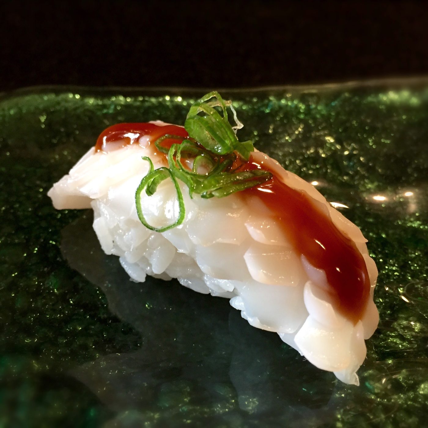 A nigiri of scored white squid brushed with dark glaze and topped with fine scallion, on a green glass plate.