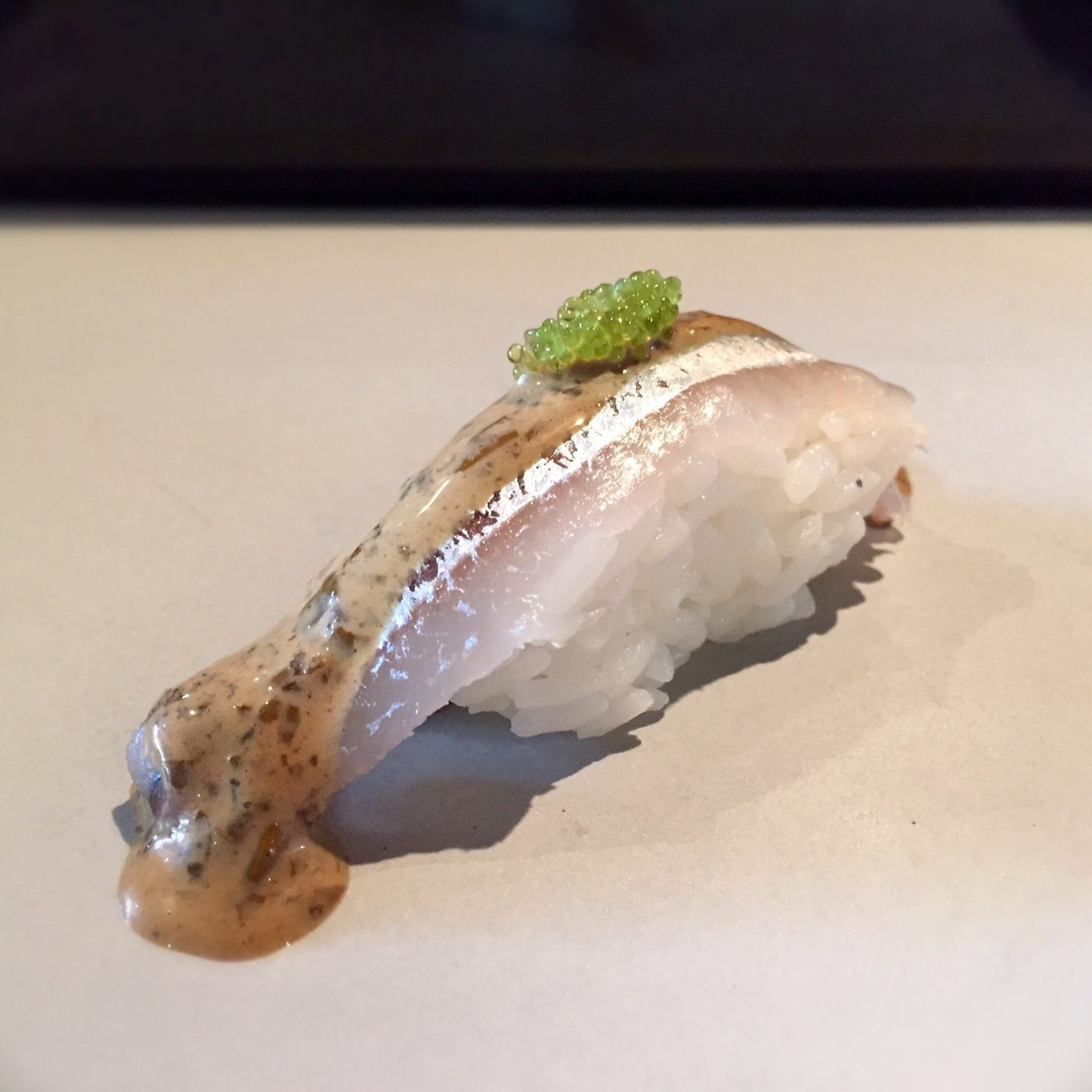 A nigiri of silver-skinned fish under a thin pale cream flecked with dark, topped with bright green roe.