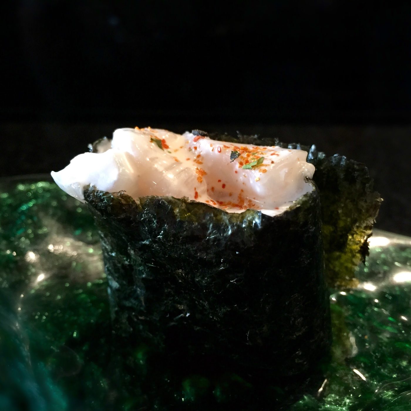 Rice walled round with nori holding a thick pale slab scored in a cross-hatch, dusted with orange-red chili and green herb, on a green glass plate.