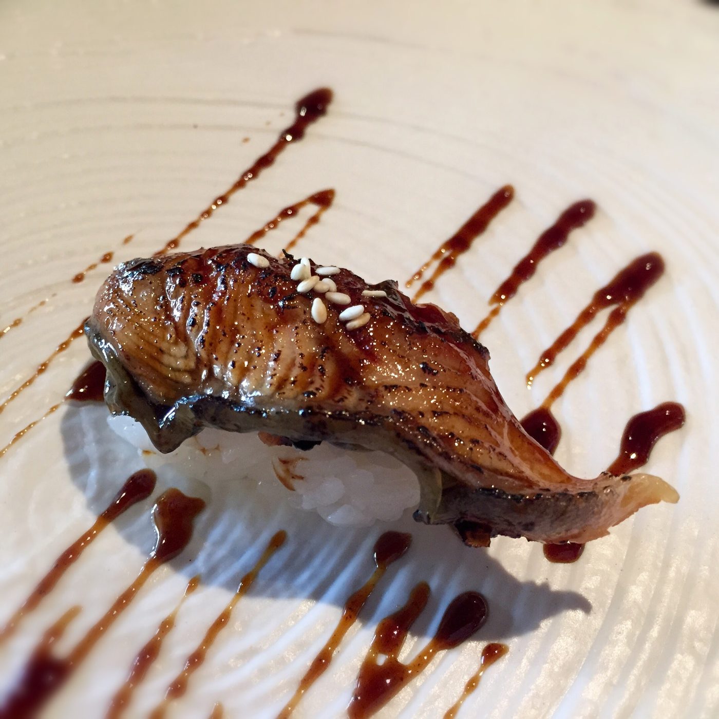 A nigiri of grilled eel lacquered dark, with glaze streaked across the plate.
