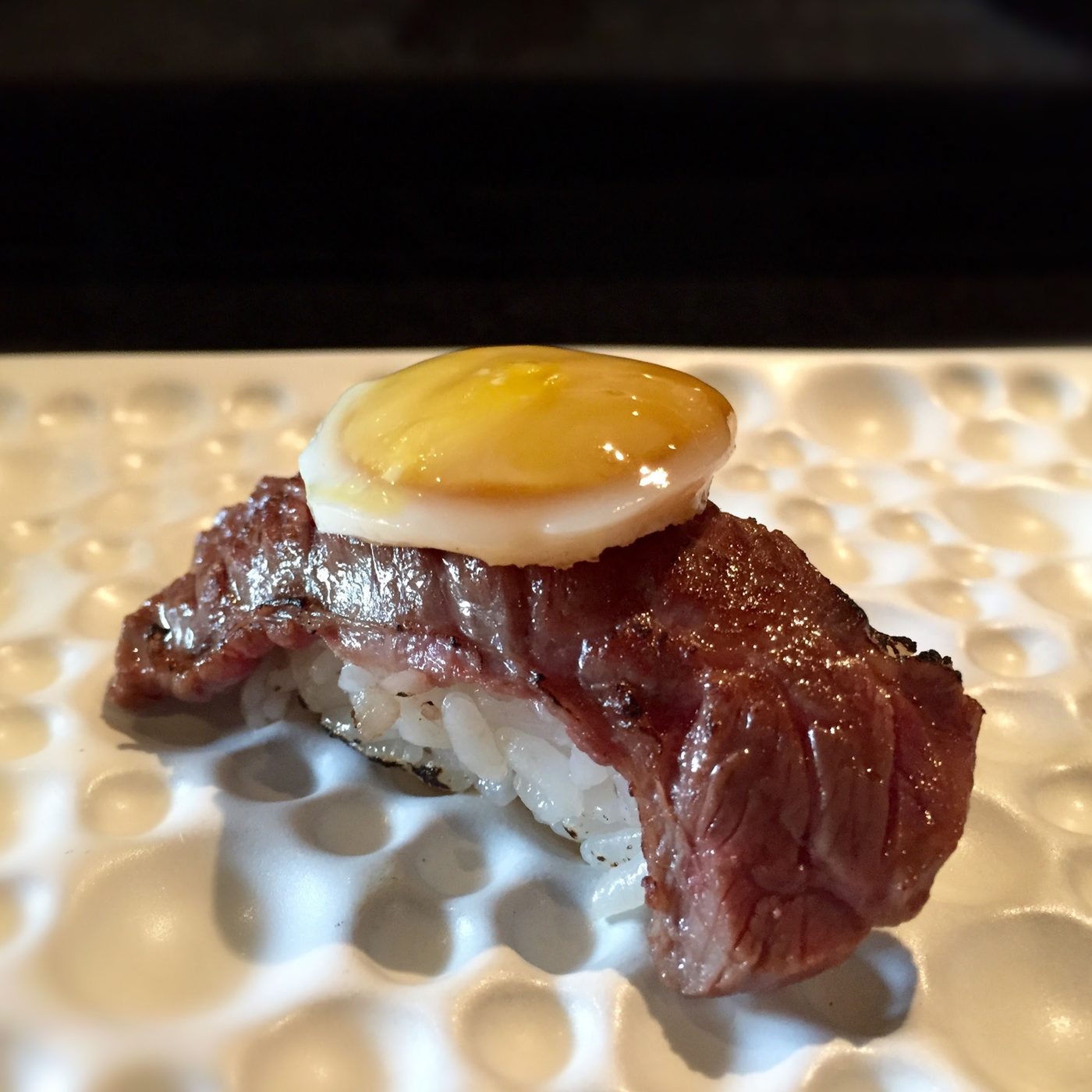 A nigiri of beef topped with a quail egg, on a dimpled white plate.