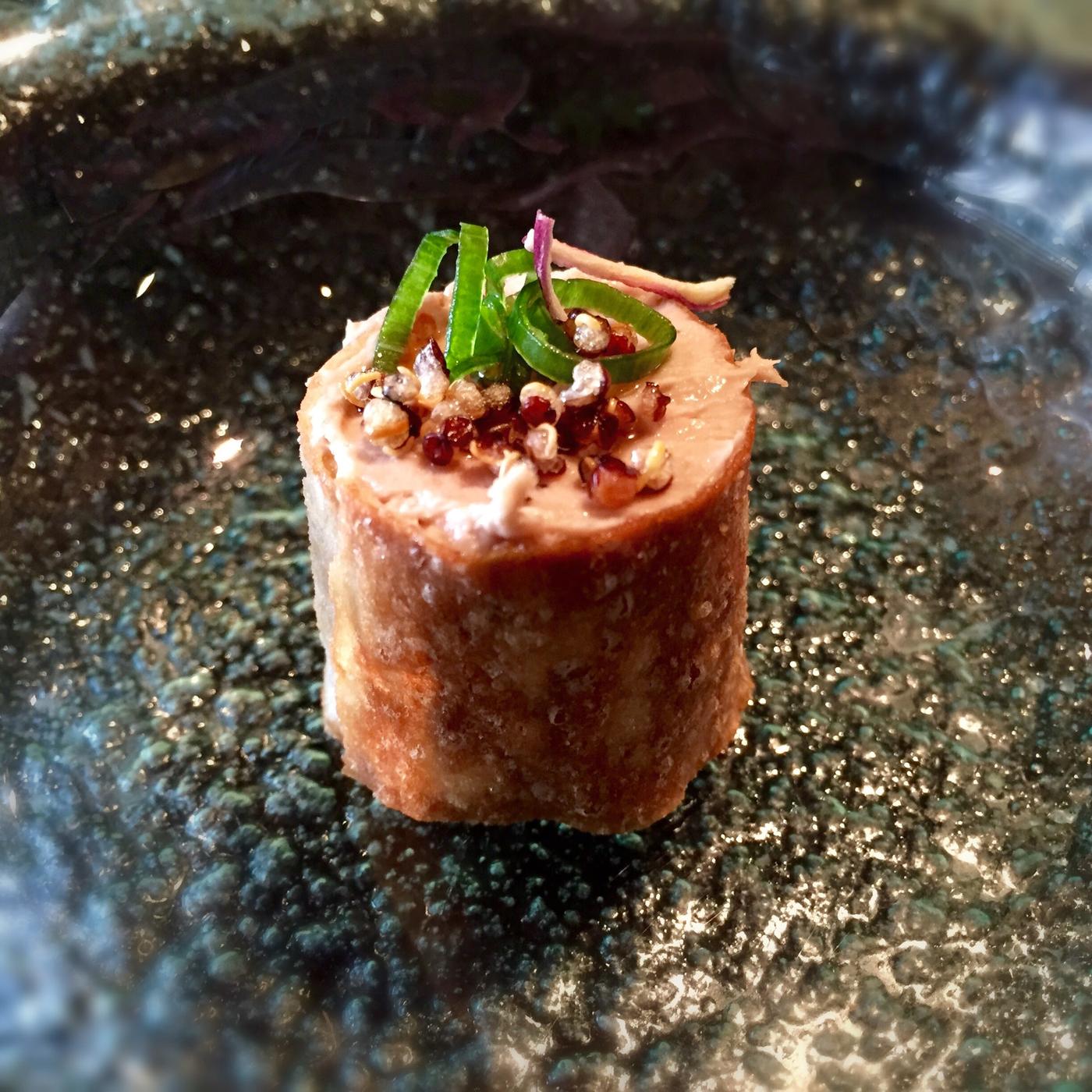 An upright cylinder in a thin blistered transparent fried wrapper, packed with pale filling, topped with popped red and white quinoa and green scallion curls.