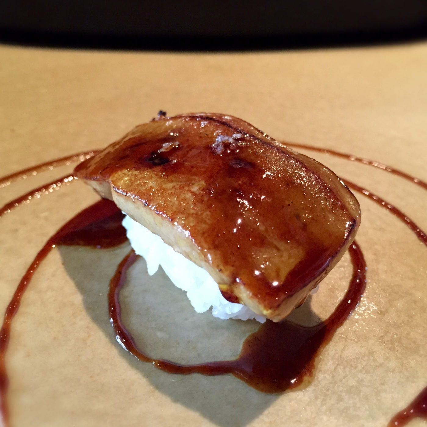 A slab of foie gras over rice under a dark glaze, with sauce swirled across the plate.