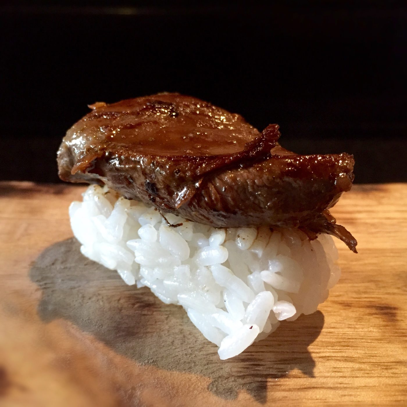 A dark glazed squab breast laid over rice on a wooden board, without a nori band.