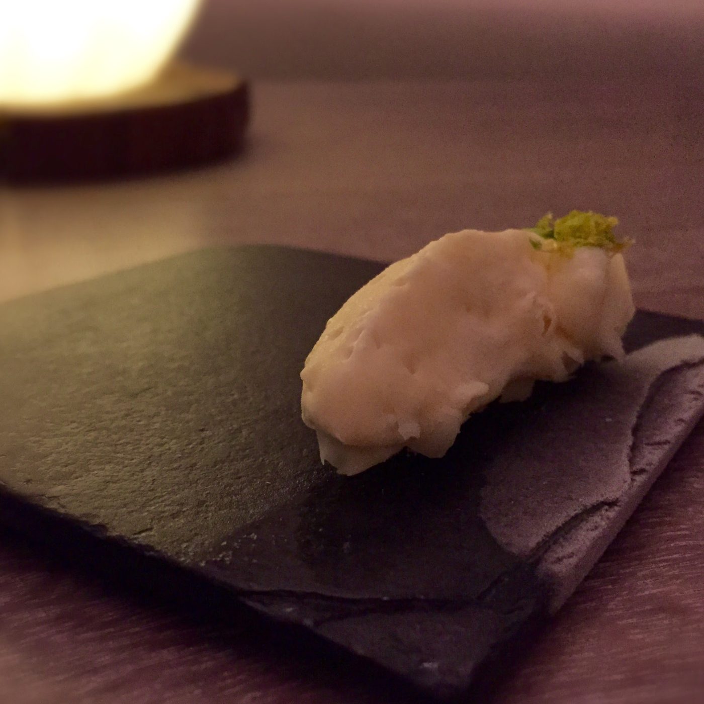 One pale quenelle on a black slate tile, its surface pitted like set ganache, with a small tuft of yellow-green zest at one end.