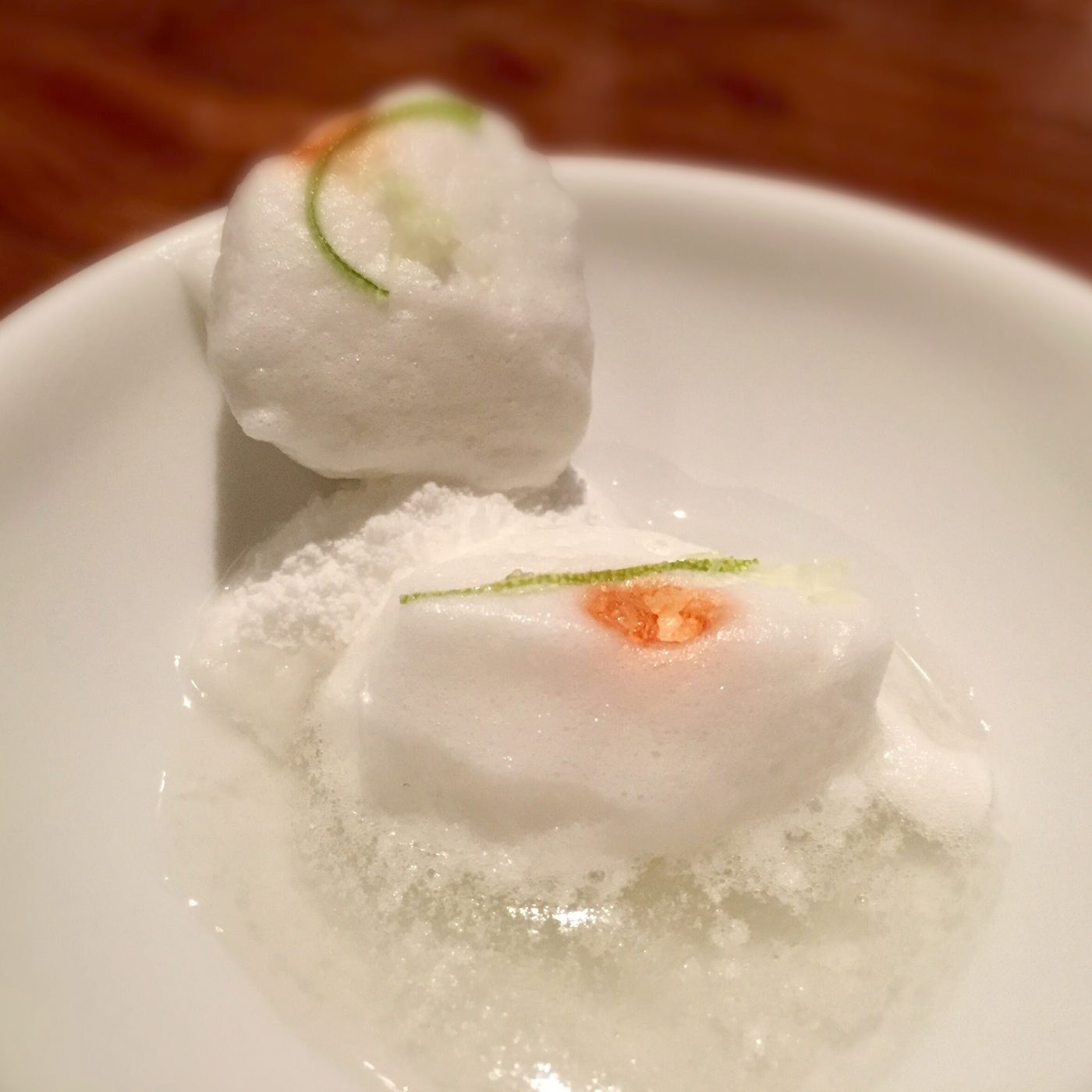 Two white aerated pieces stacked on a white plate, each with a thread of citrus zest laid across it and a single orange dot on the lower one.