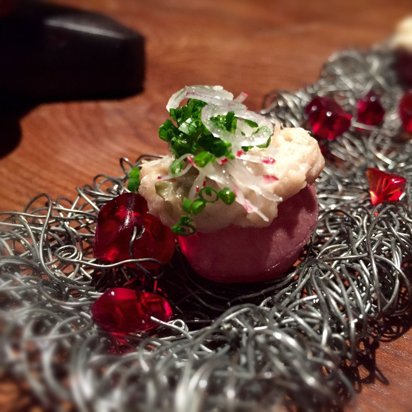 A tangled wire nest strung with red glass beads, holding a deep magenta disc topped with pale shredded meat, shaved radish and chopped chive.
