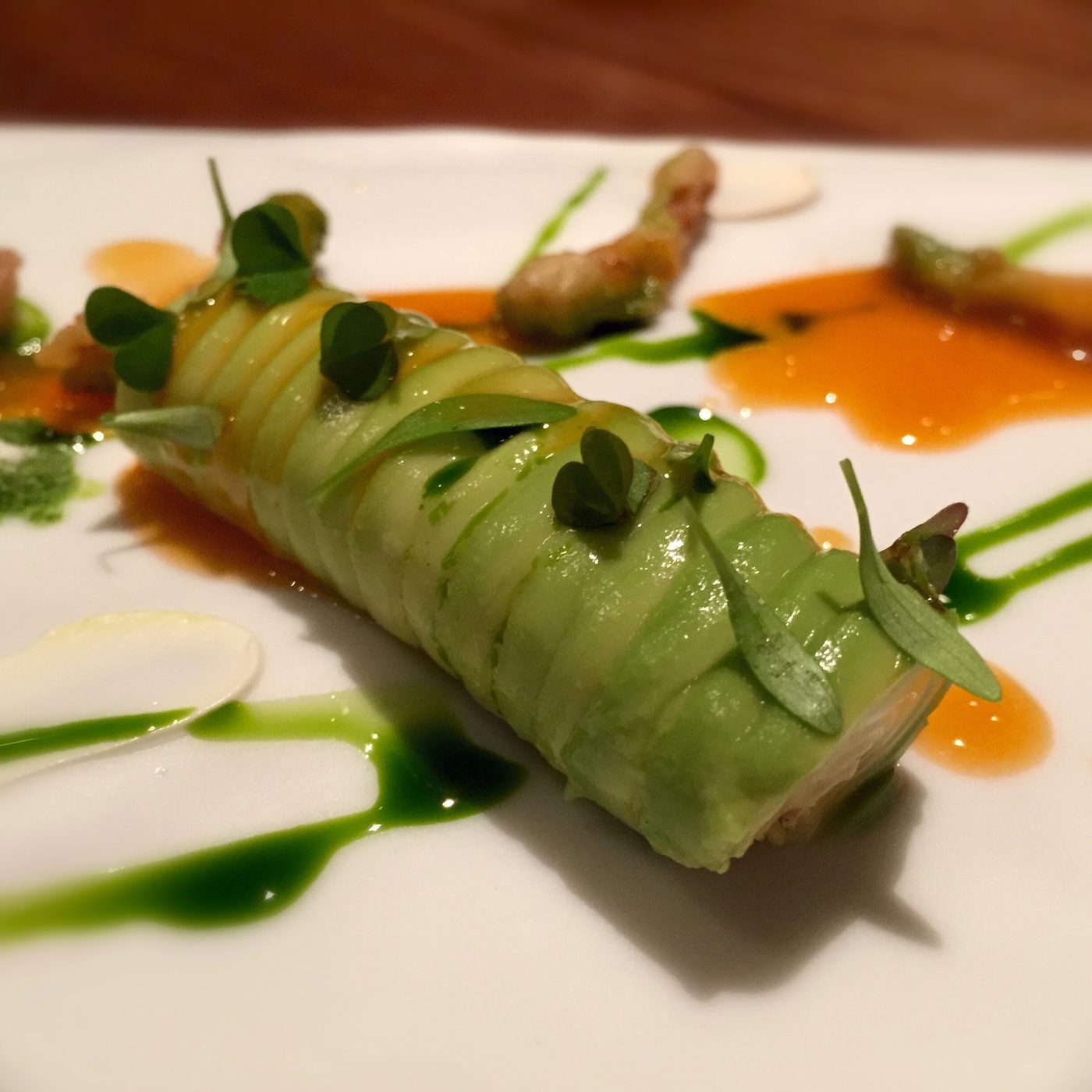 A cylinder wrapped in overlapping avocado slices with micro leaves pressed along it, an orange sauce and green oil run across the plate, small fried pieces alongside.