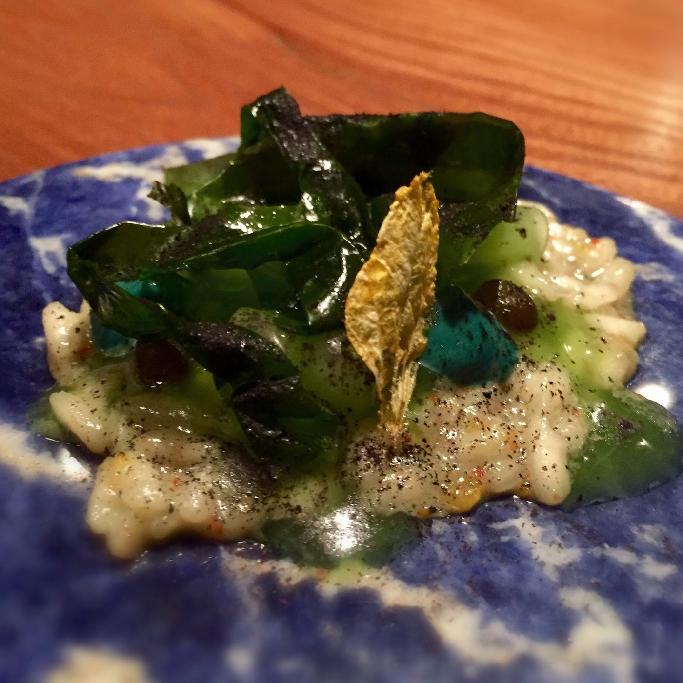 An indigo-and-white mottled plate holding loose creamy rice dusted with fine black-green powder, heaped with glossy dark seaweed leaves and translucent pale-green cucumber.