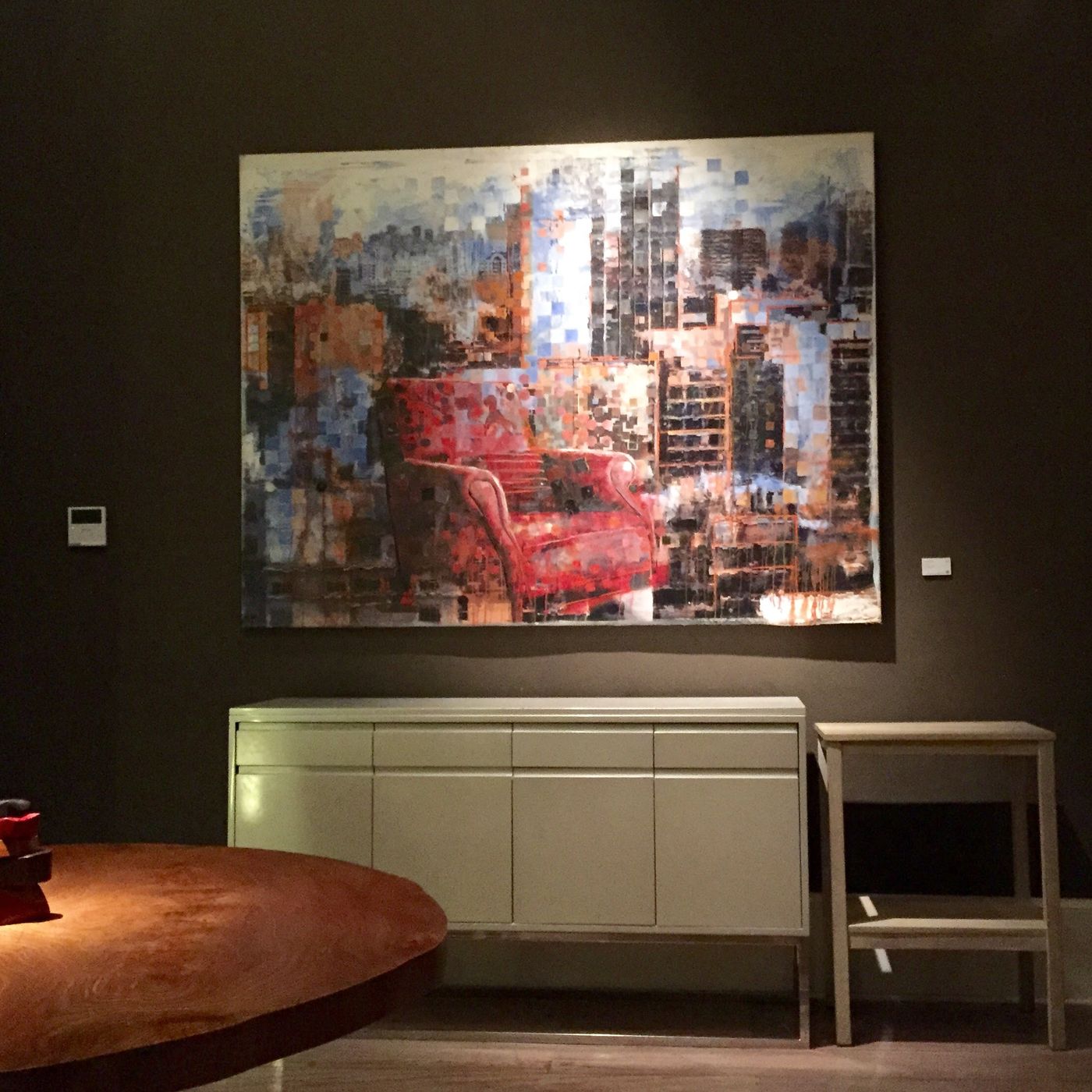 A large painting of a red armchair dissolving into a pixelated cityscape, hung above a pale sideboard, with the edge of a round wooden table in the foreground.
