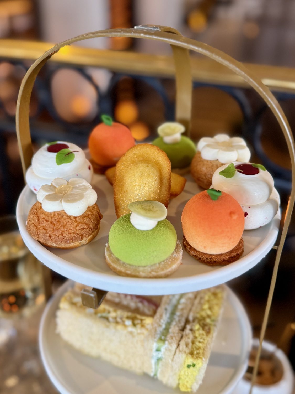 An afternoon tea tier of desserts — Tiramisu Choux, Orange Mousse, Raspberry Pavlova, Matcha Entremet and Madeleines — with finger sandwiches on the level below