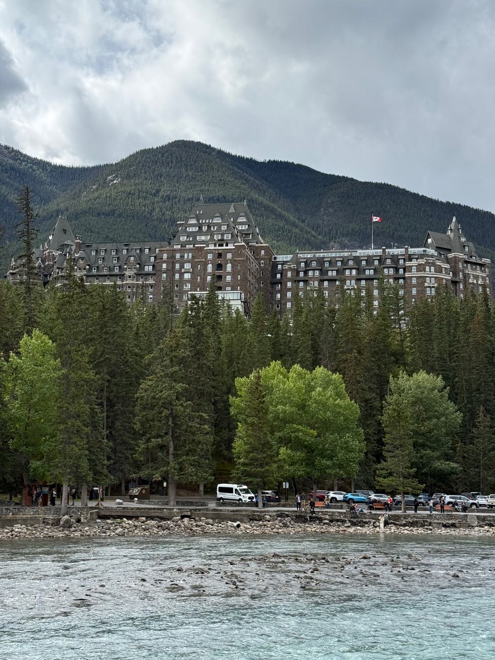 The Banff Springs Hotel seen from the bank of the Bow River, rising above the forest on the far side under grey cloud