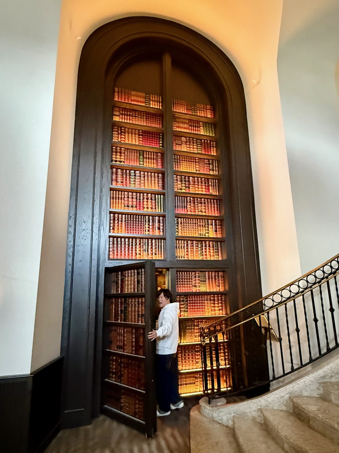 A floor-to-ceiling bookcase filling a stone arch beside a staircase, standing open as a door into the next room, with a figure at the opening
