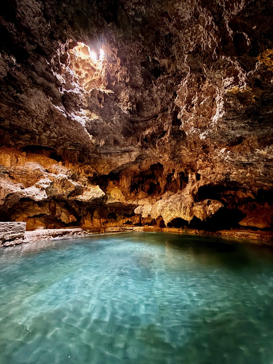 A turquoise hot spring pool inside a cave, lit from an opening in the rock vault above