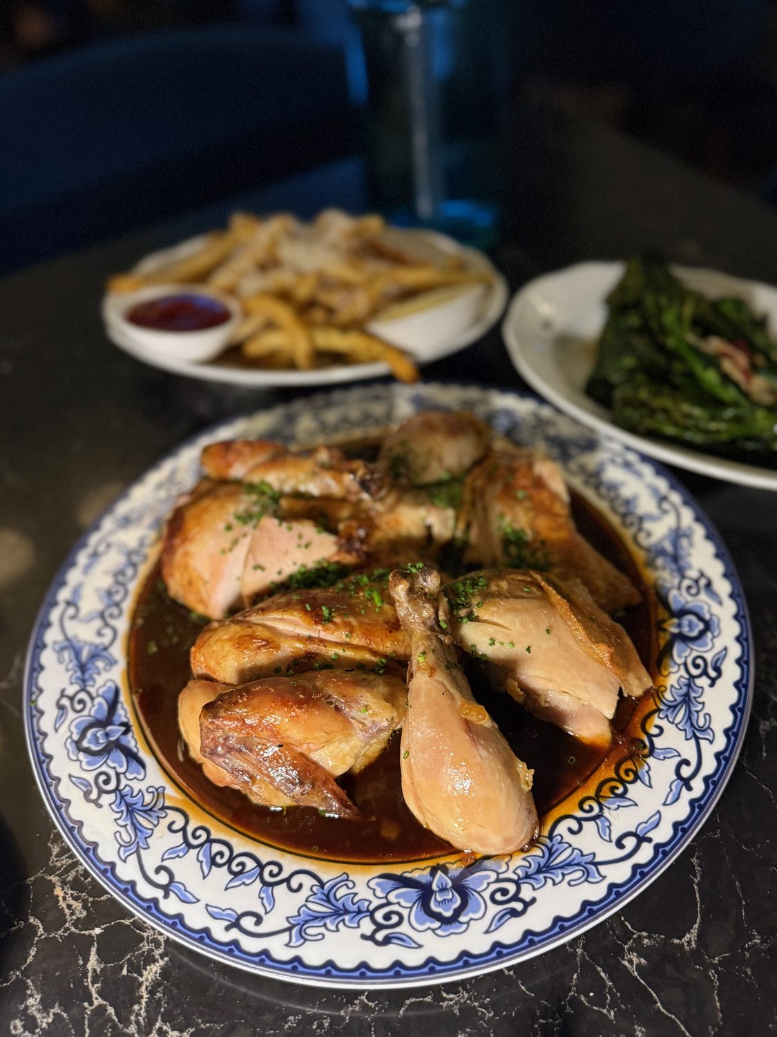 A whole rotisserie chicken carved onto a blue and white porcelain platter with a scrolling floral border, sitting in a pool of dark glossy jus with chives, truffle fries and charred broccolini behind it