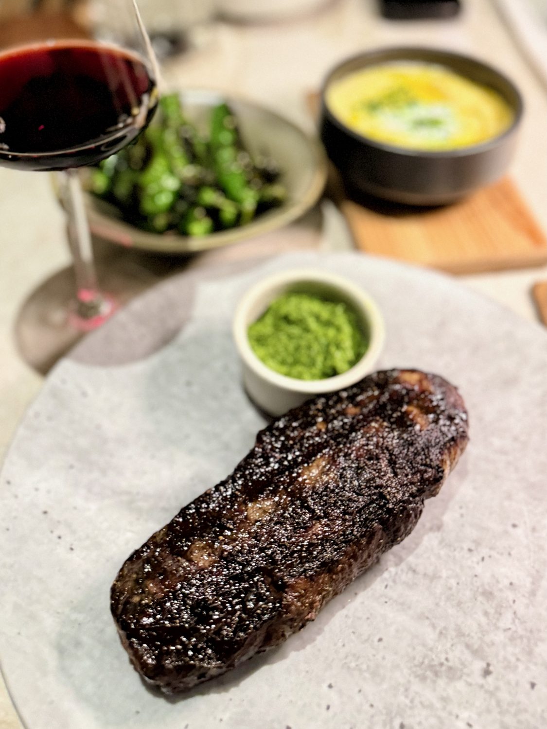 A seared wagyu striploin on a pale stone plate with a ramekin of green chimichurri, a glass of red wine and two side dishes behind it