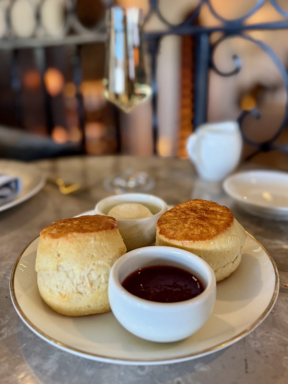 Two warm scones on a gold-rimmed plate with small pots of clotted cream and strawberry preserve beside them
