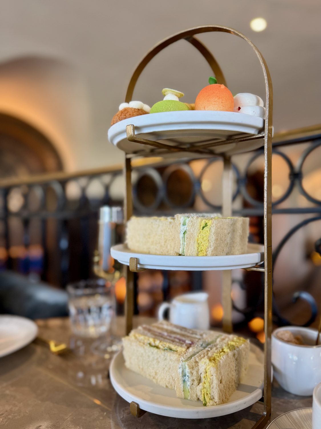 A three-tier afternoon tea stand on a marble table, desserts on the top tier and finger sandwiches on the two below, with ironwork and a lit bottle niche behind