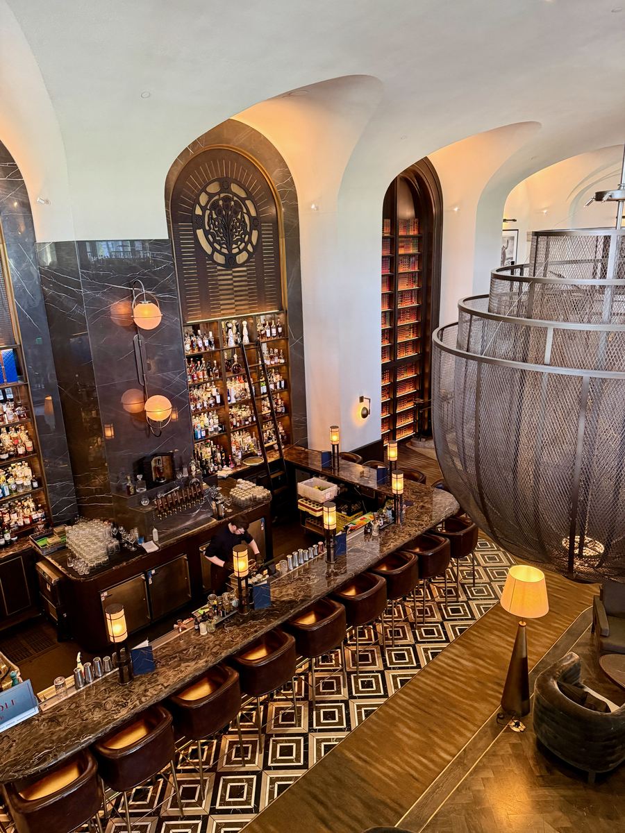 The Rundle Bar seen from the floor above: a long marble counter, bottles stacked up an arched niche, and a black and white geometric floor under vaulted ceilings