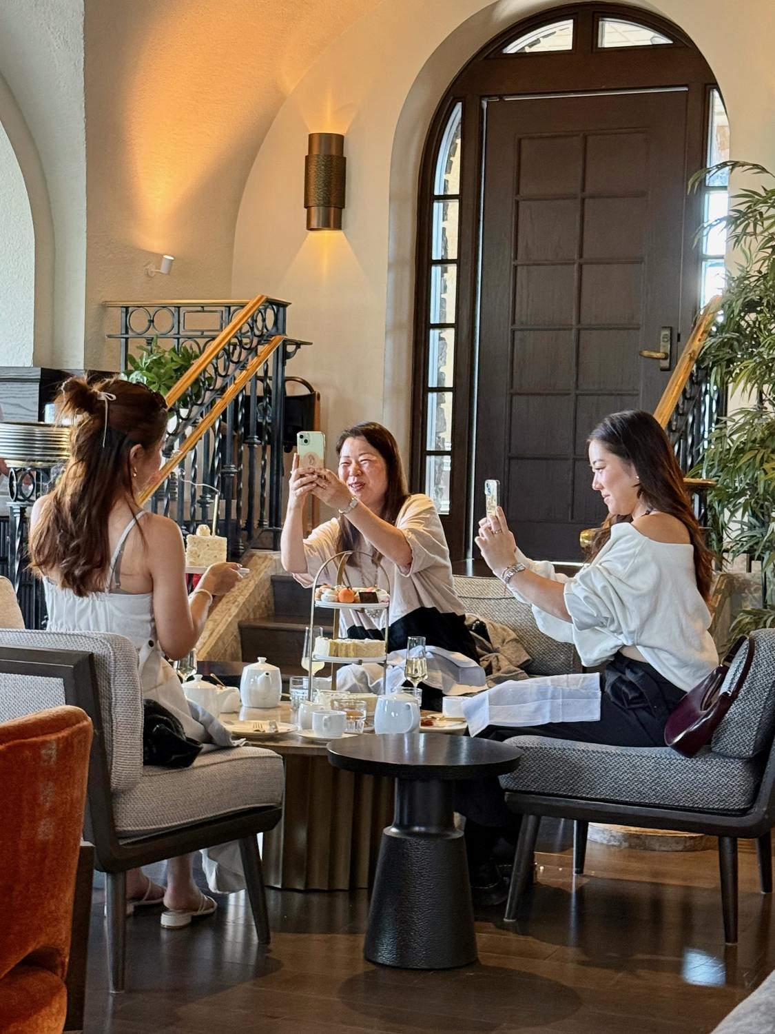Guests at a table photographing their tiered afternoon tea stand before eating, one holding a tall slice of cake, beneath an arched wooden door