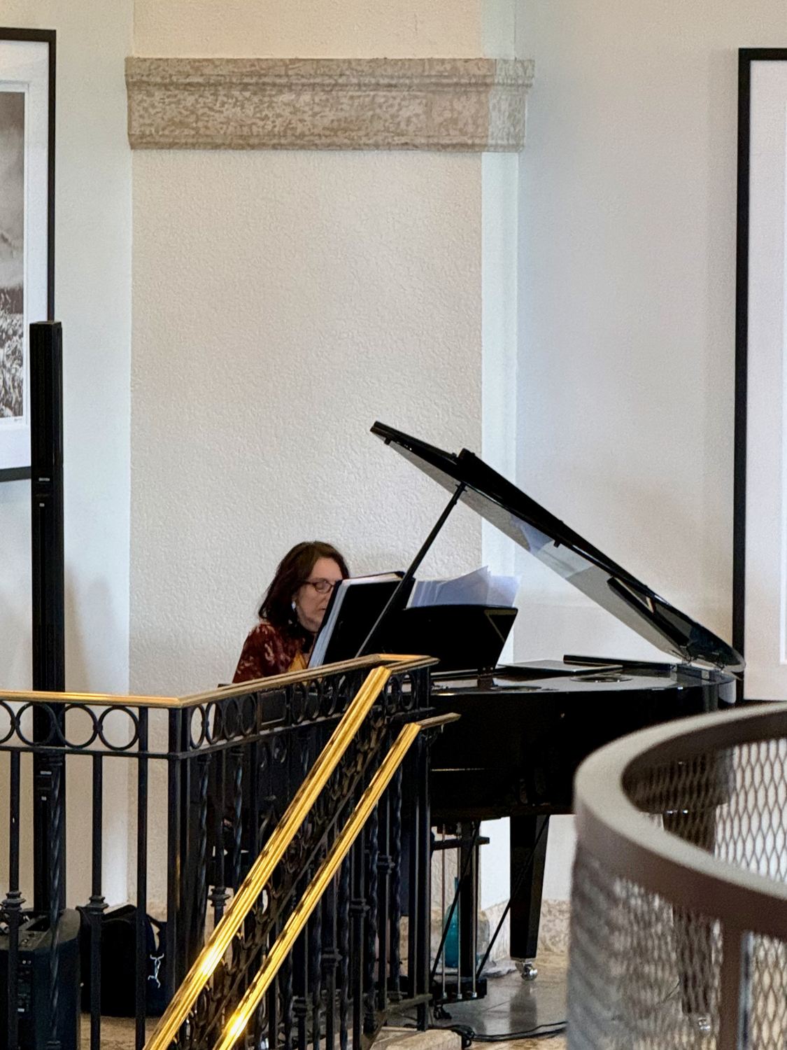 A grand piano being played on the tea floor, seen past a gilded ironwork balustrade