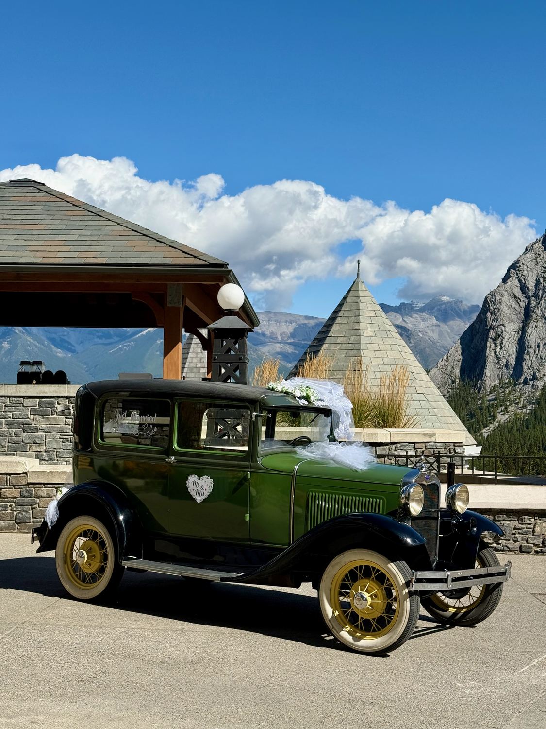 A vintage green and black sedan with yellow spoked wheels and whitewall tires parked on the hotel terrace, dressed with white tulle and flowers and Just Married lettered across the rear window, mountains behind