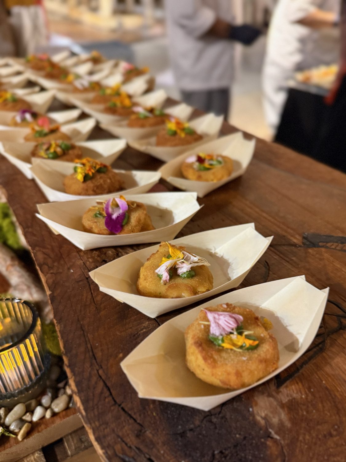 Rows of geoduck croquettes in paper boats, each topped with green pesto and edible flowers, lined up along a timber counter