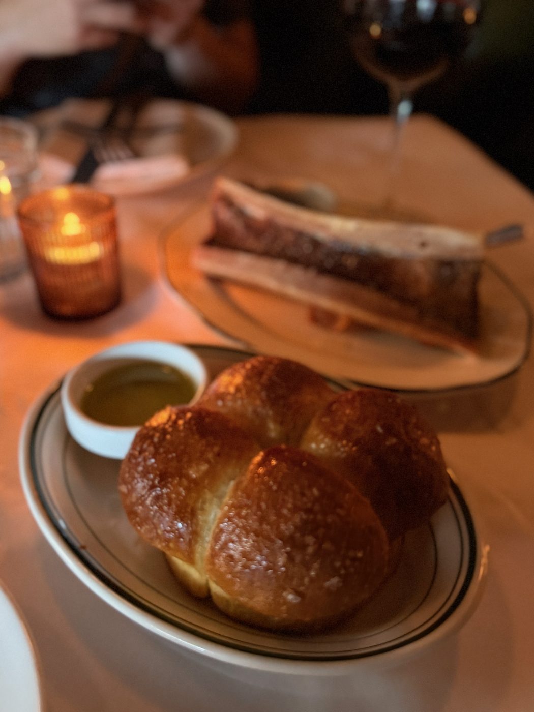 A salted potato focaccia roll on a plate beside a ramekin of olive oil, with a split marrow bone and a glass of red wine on the table behind