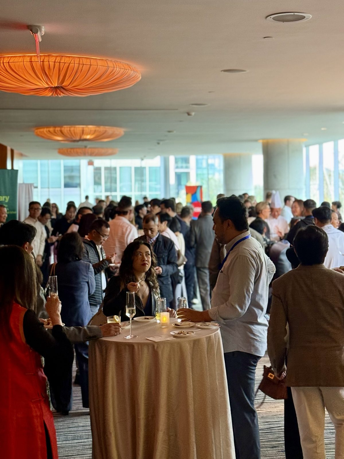 A crowded reception, with guests at a tall cocktail table holding flutes of sparkling wine beneath orange pendant lamps
