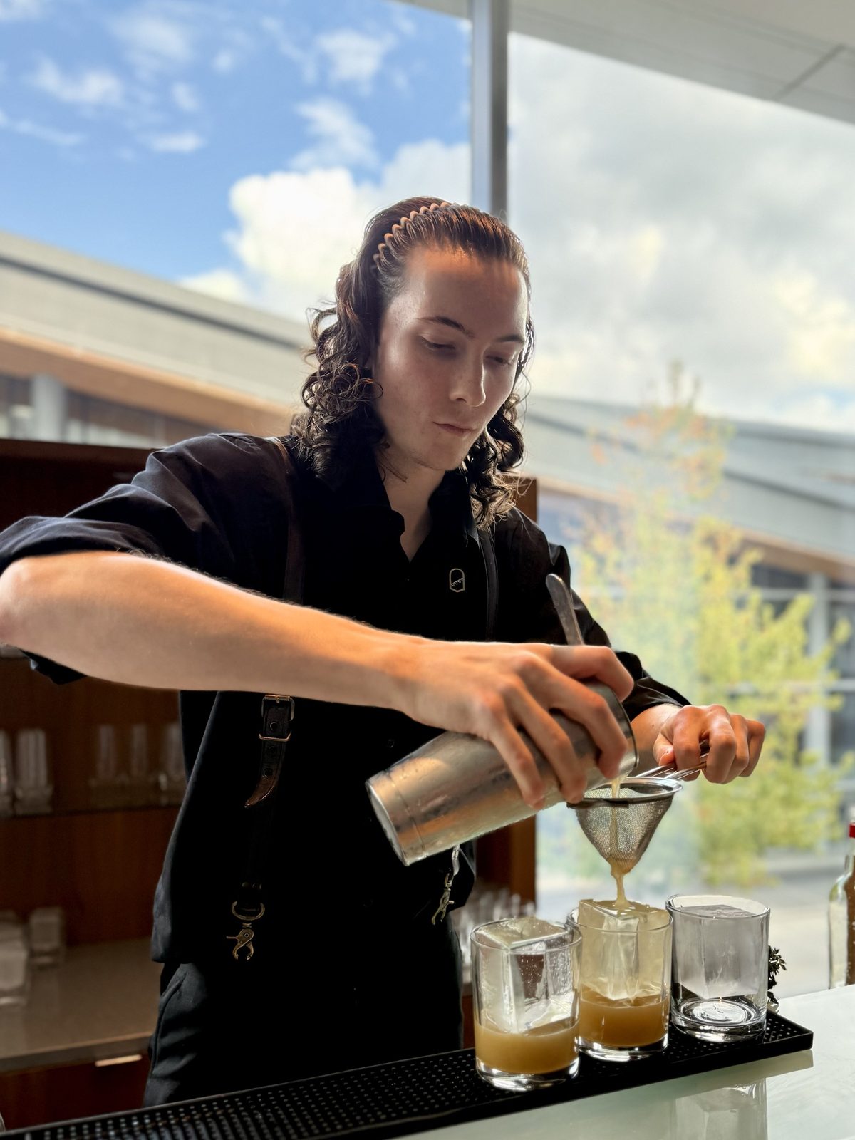 A bartender double-straining a pale gold cocktail over large clear ice cubes in rocks glasses