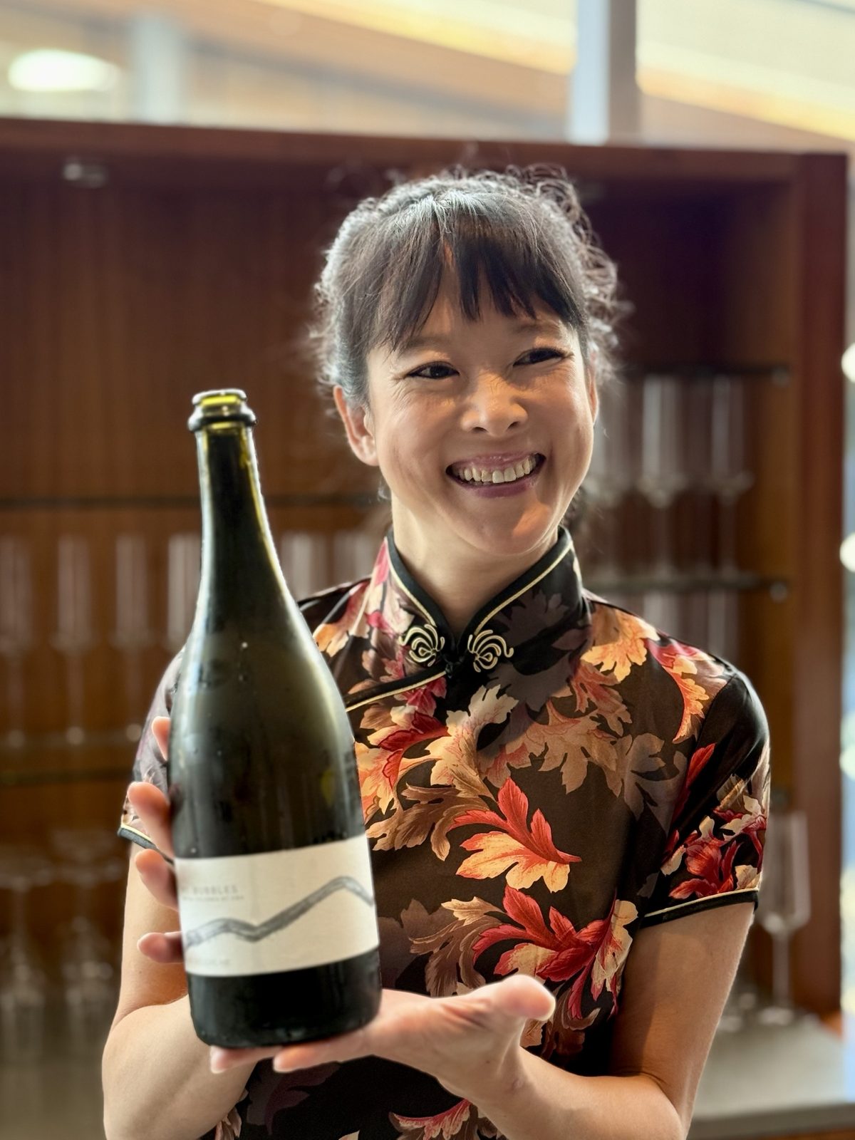 A sommelier in a floral qipao smiling and holding a bottle of BC sparkling wine