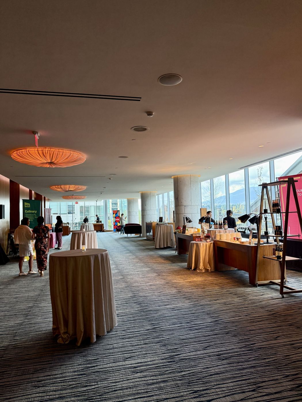 An empty reception area with tall draped cocktail tables, orange fabric pendant lamps and floor-to-ceiling windows looking toward the North Shore mountains