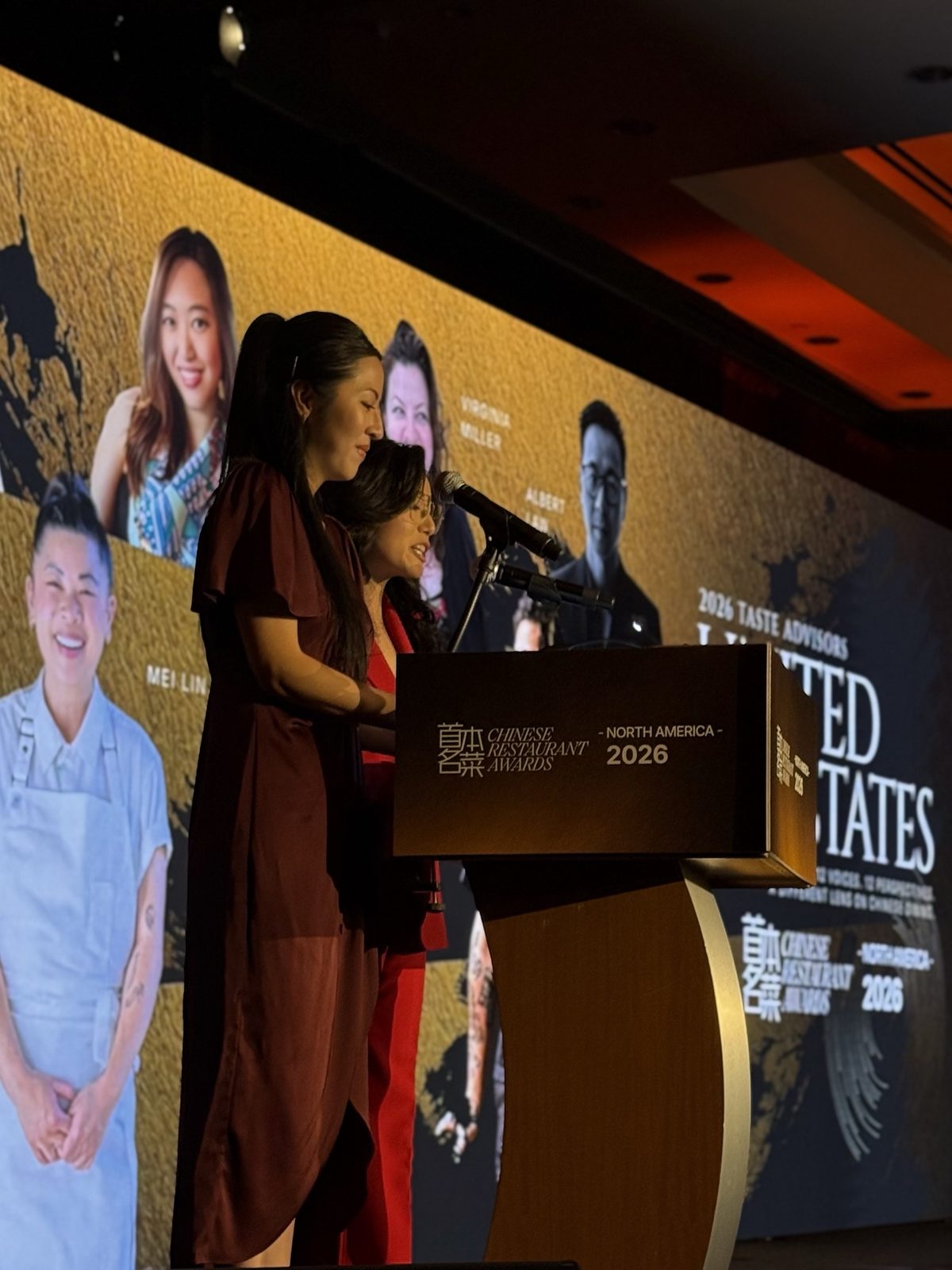 Two emcees at a podium in front of a screen showing portraits of this year’s Taste Advisors