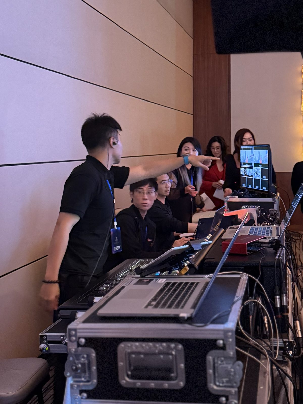 A production crew with earpieces at a desk of laptops and a video switcher, with presenters waiting behind