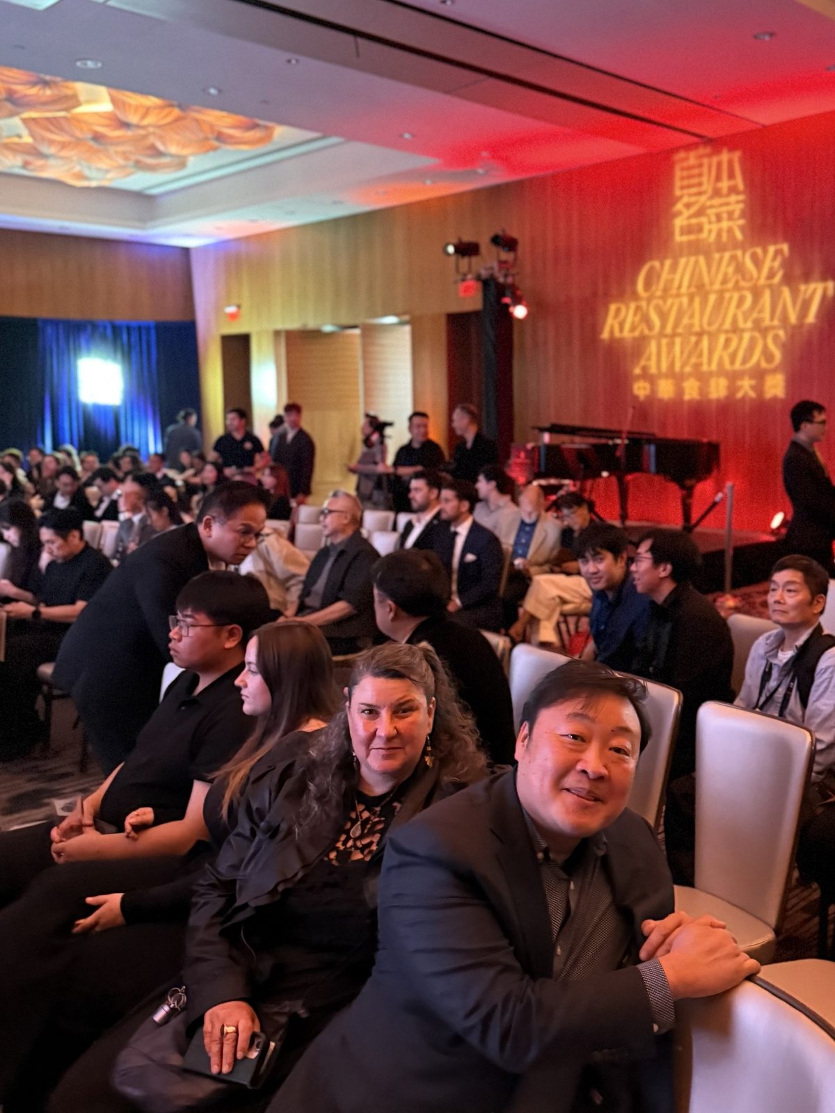 A ballroom set as an auditorium, with guests in rows of pale chairs facing a stage washed in red light, the Awards’ name projected in gold and a grand piano to one side