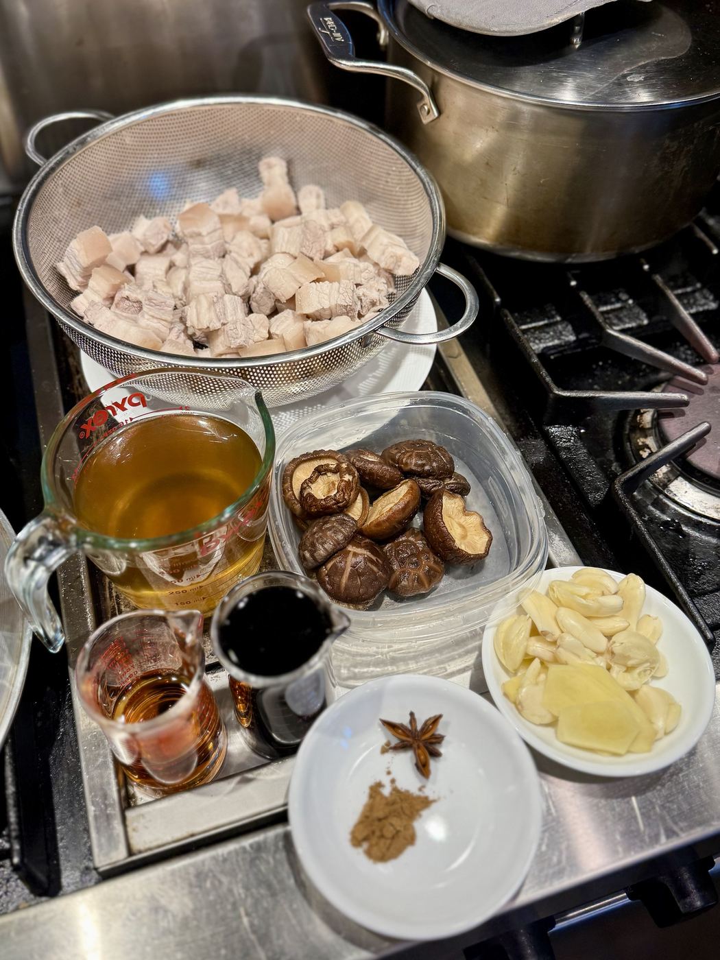 Prepped ingredients by the stove: blanched pork belly in a colander, mushroom soaking liquid, Shaoxing wine and soy in measuring cups, rehydrated shiitake, garlic and ginger, and star anise with five-spice in a small dish
