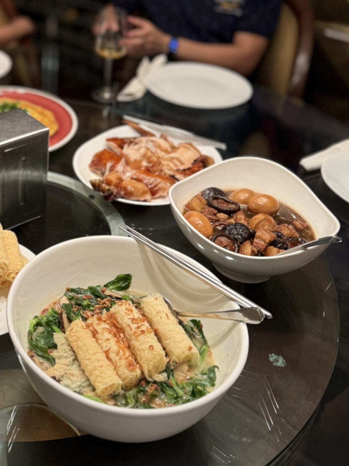 Hong bah with whole eggs in a white bowl on a lazy Susan, beside roast meat and garlic pea tips with bean curd rolls