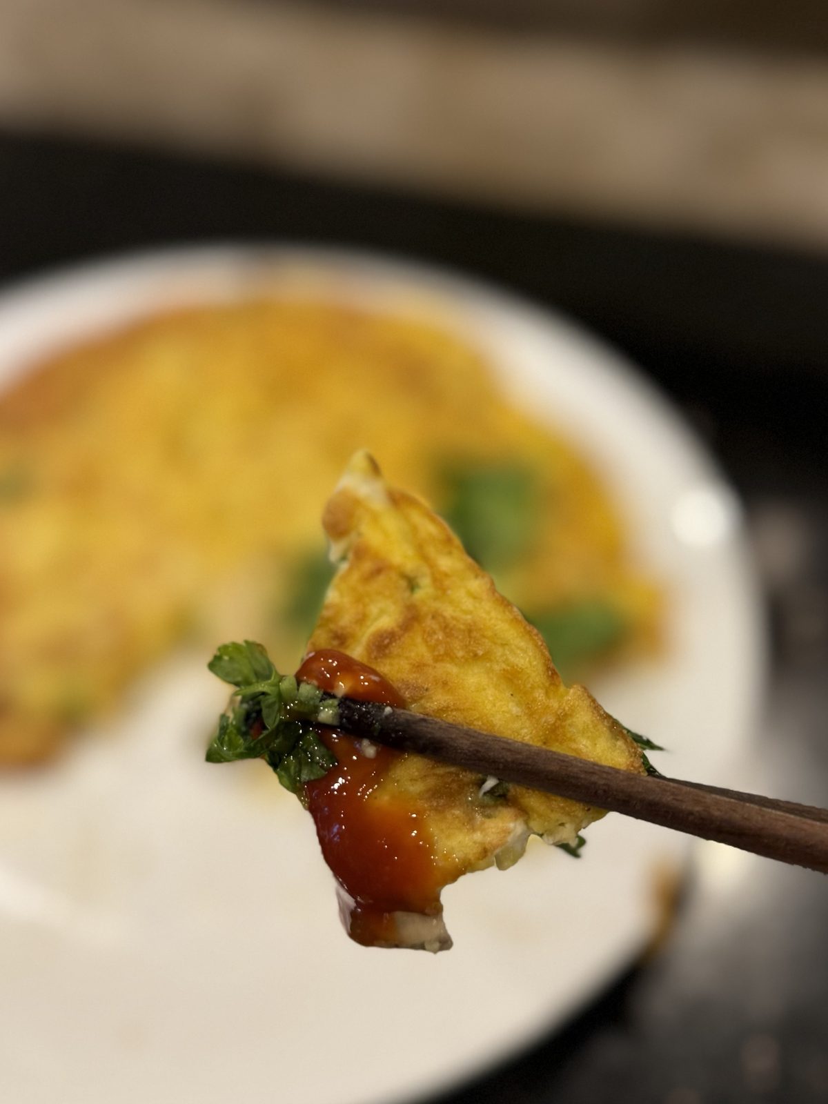 A piece of oyster omelette held in chopsticks, dipped in ketchup