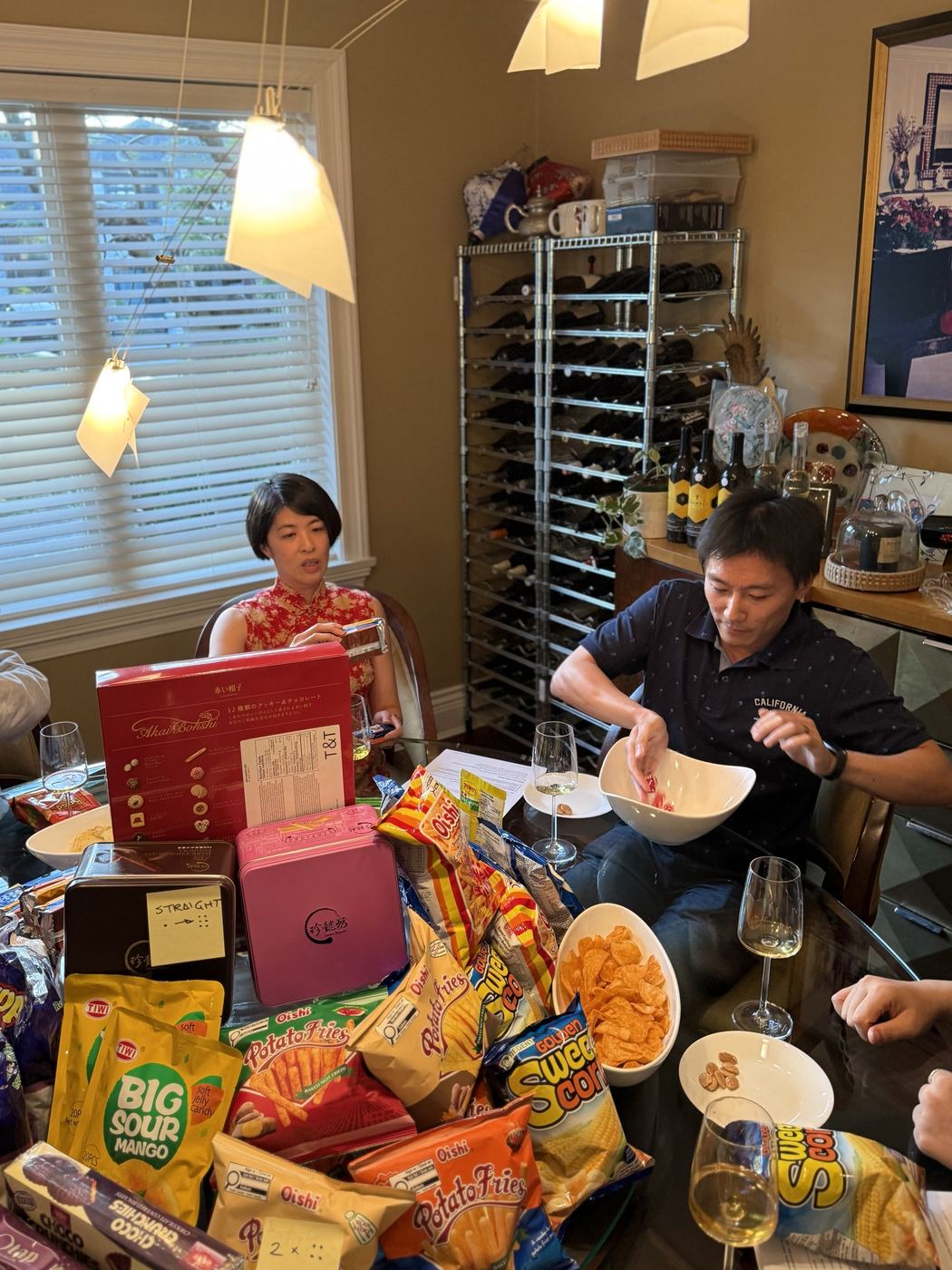 A guest rolls dice into a white bowl at a table crowded with snack bags and prize tins labelled with sticky notes