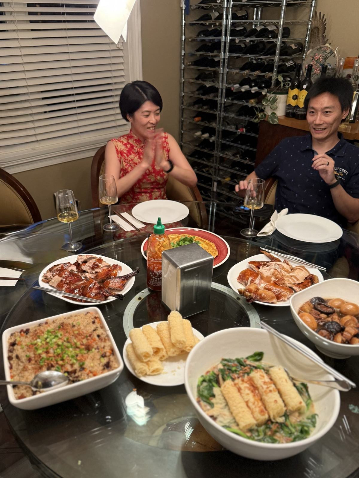 A round table set with roast duck, soy chicken, braised pork belly with eggs, an omelette, sticky rice and pea tips in soup, two guests laughing behind
